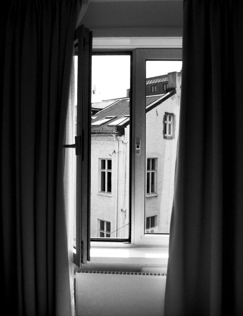 A monochrome view of an exterior building through an open window with curtains.